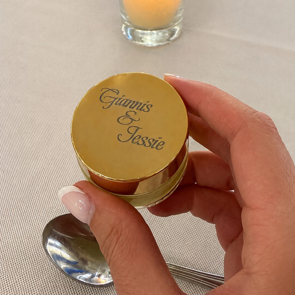 Gold jar cap held in hand, engraved with two first names, at a table setting with a lit candle